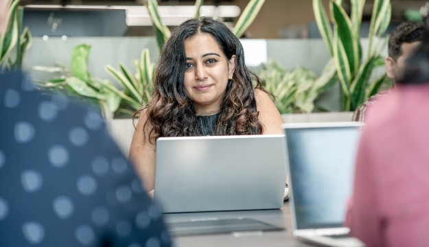 Person seated at a meeting table using a laptop in a modern office environment, with colleagues partially visible in the foreground and indoor plants incorporated into the workspace behind