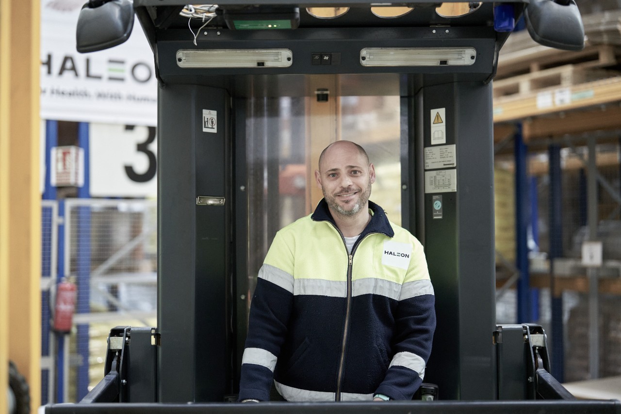 Person wearing a Haleon-branded high-visibility jacket standing inside an industrial warehouse environment, positioned within a forklift cab, with warehouse storage racking, equipment, and Haleon signage visible in the background.