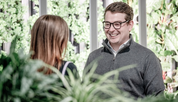Two people standing and talking in a modern office environment with indoor plants and a vertical green wall feature in the background, viewed through foreground planting within a collaborative workspace