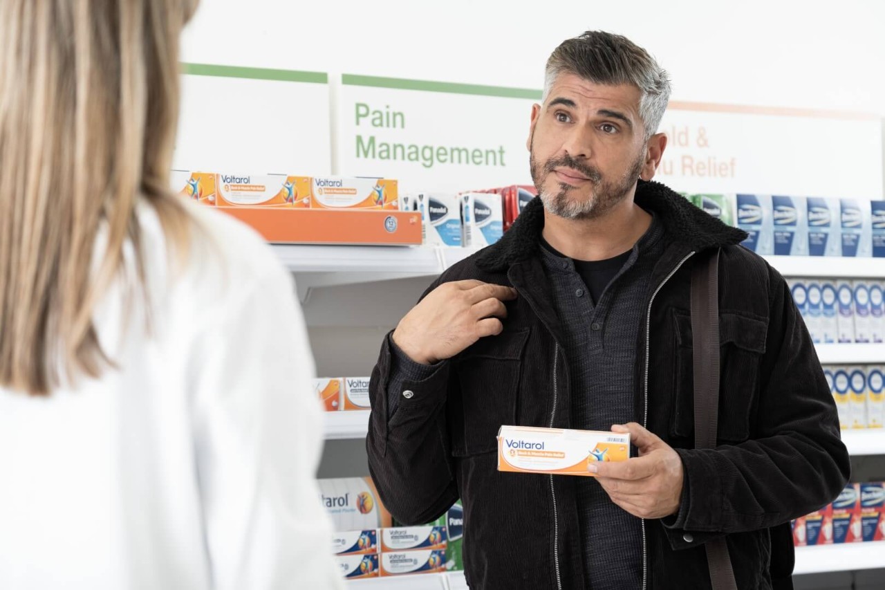 A person talking to a healthcare professional in a pharmacy holding a tube of Voltaren in one hand while pointing to his shoulder with the other 