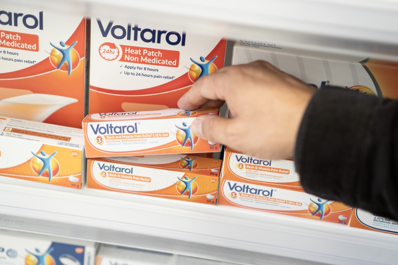 Hand selecting Voltarol gel for back and muscle pain relief from a shelf filled with various Voltarol products