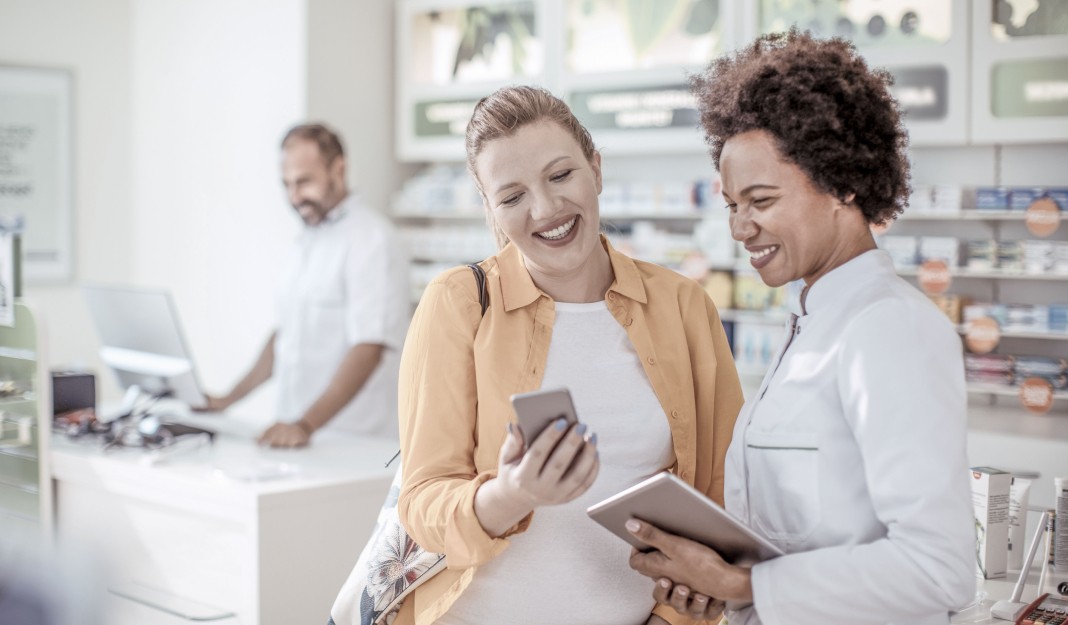 Two smiling women, one in pharmacist attire, looking at a phone screen, while another pharmacist in the background uses a computer