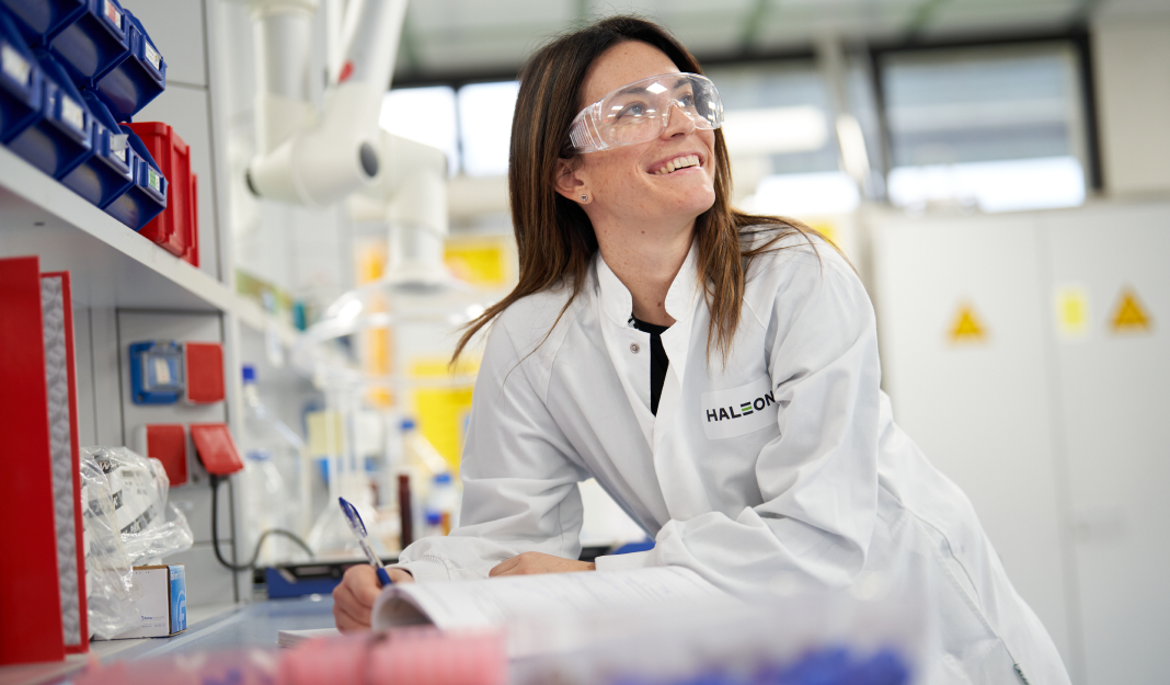A woman in a science lab wearing a white lab coat and protective glasses smiling while looking to the side  