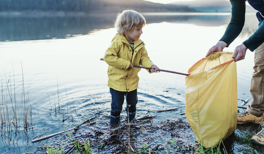 Young boy in yellow raincoat collects items from lake with father's help, using shovel handle and yellow bin bag.