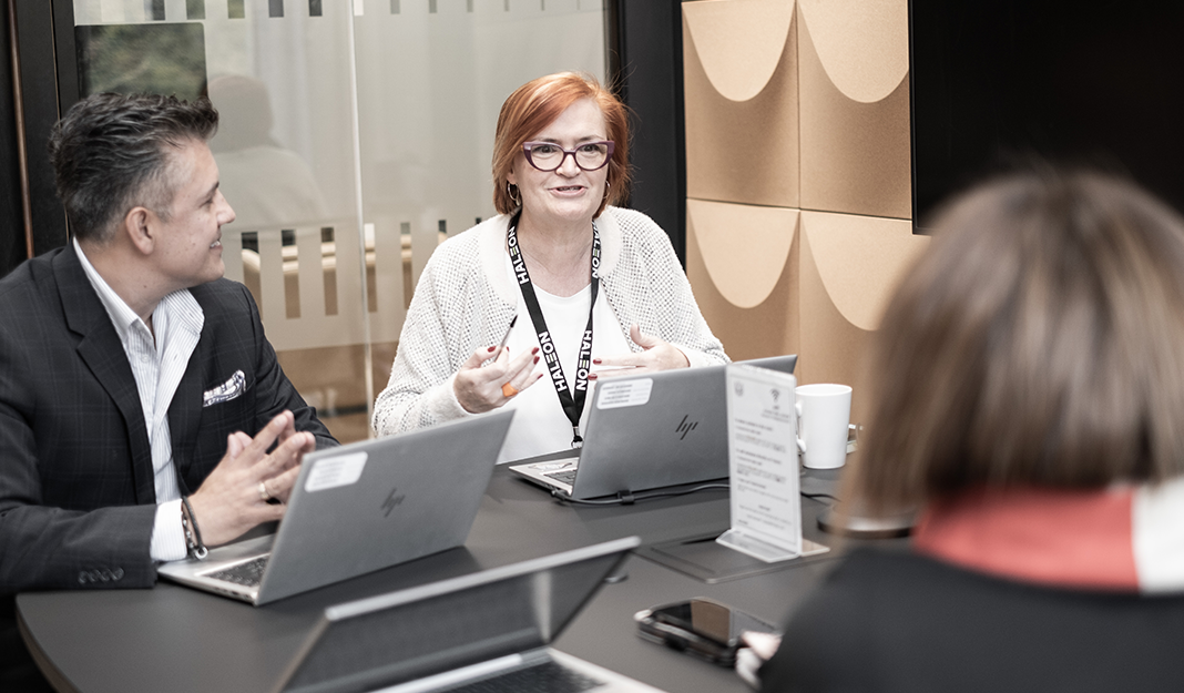 Three people seated around a table in a meeting room, using laptops and discussing information, with notebooks and documents visible
