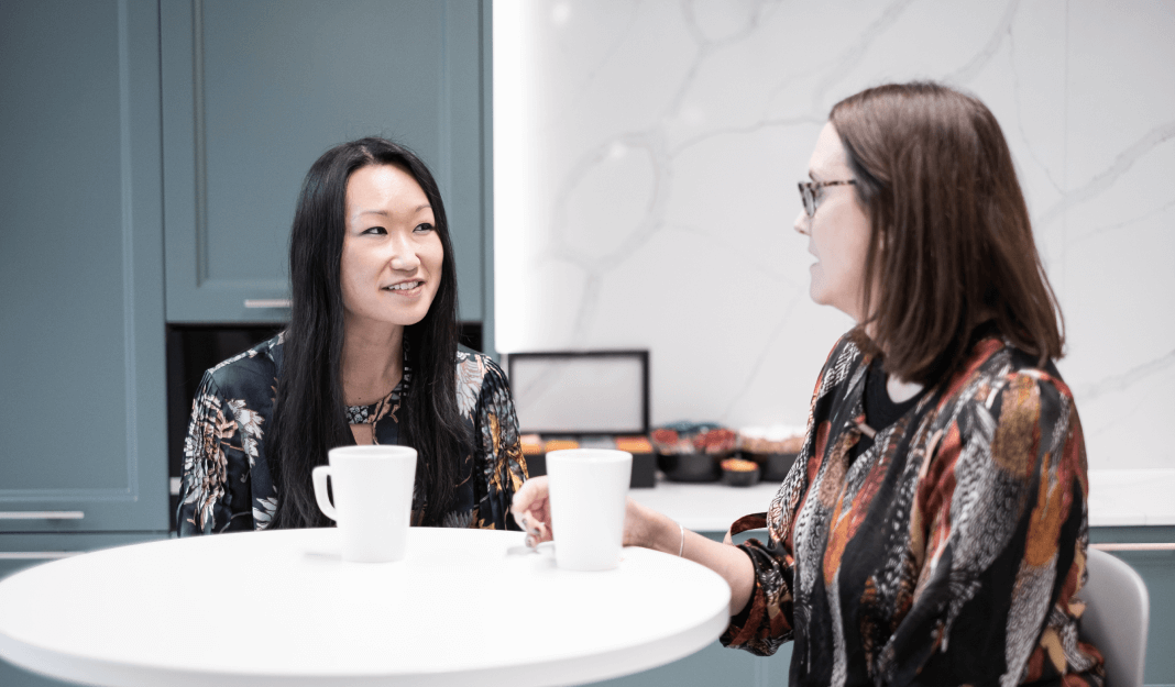 Two colleagues sitting at a small table, talking over coffee in a modern office kitchen area.