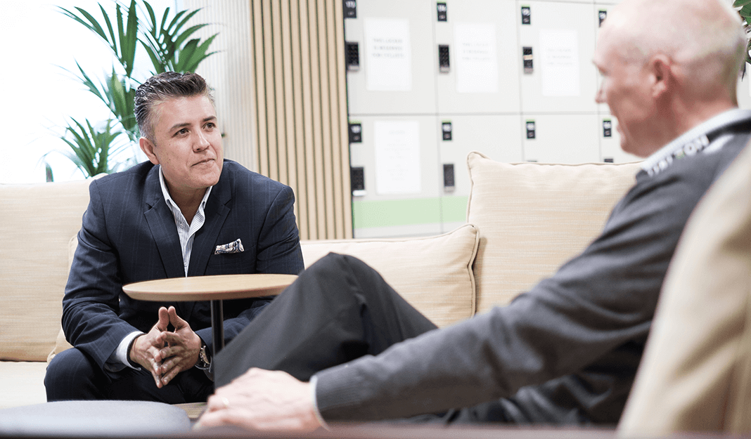 Two people seated and talking in a modern office lounge area, with soft seating, indoor plants, and lockers visible in the background